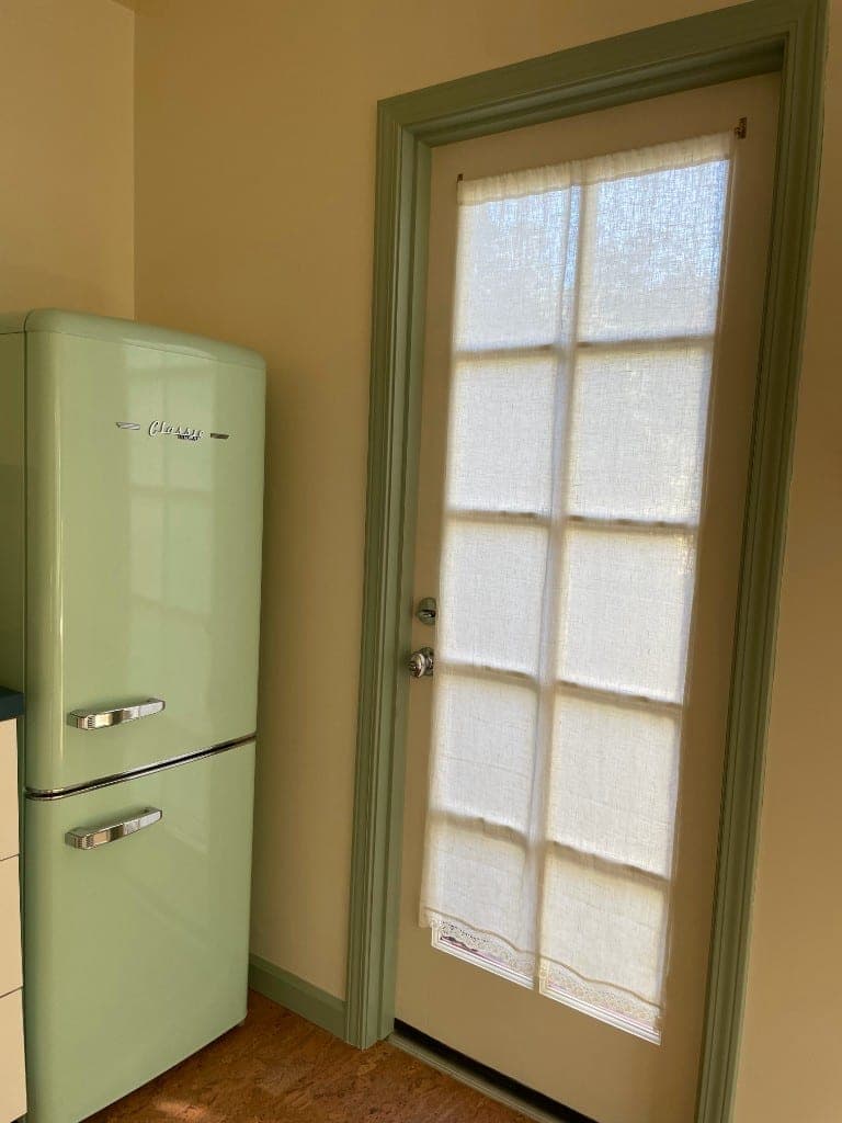 Mint vintage Classic refrigerator with chrome handles beside a sage-trimmed door and lace curtain